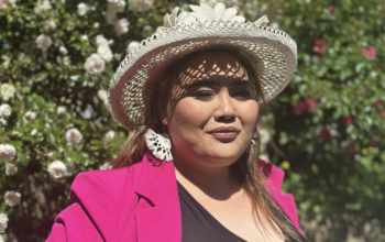 A women wearing a bight pink jacket and a sunhat. In the background is a rose garden.